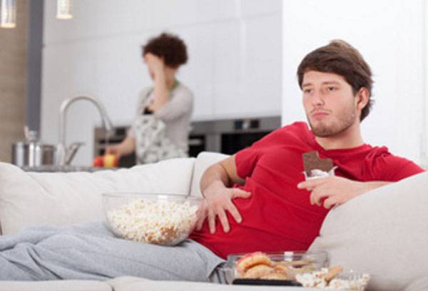 Der Ehemann liegt mit Popcorn auf dem Sofa. Im Hintergrund fasst sich die Frau an den Kopf.